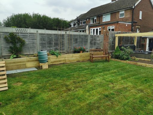 A green garden with wooden decking, a bench, and a grey fence surrounding a house.