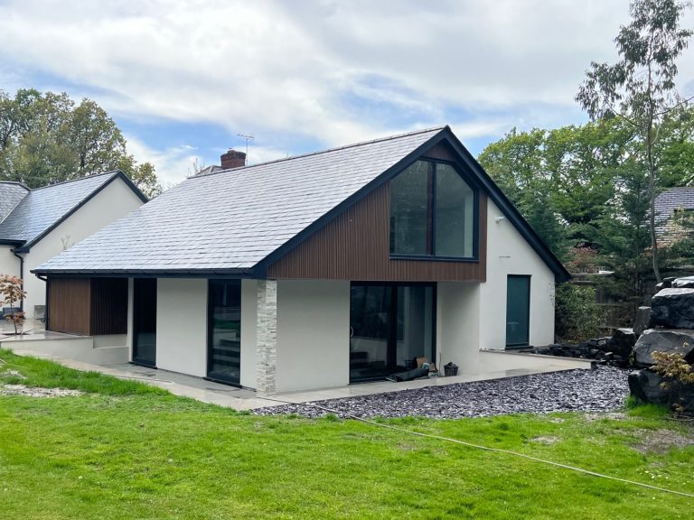 Small raised grey wooden deck in a garden with a round glass-top table and two wicker chairs. The deck sits beside grey patio tiles and is b